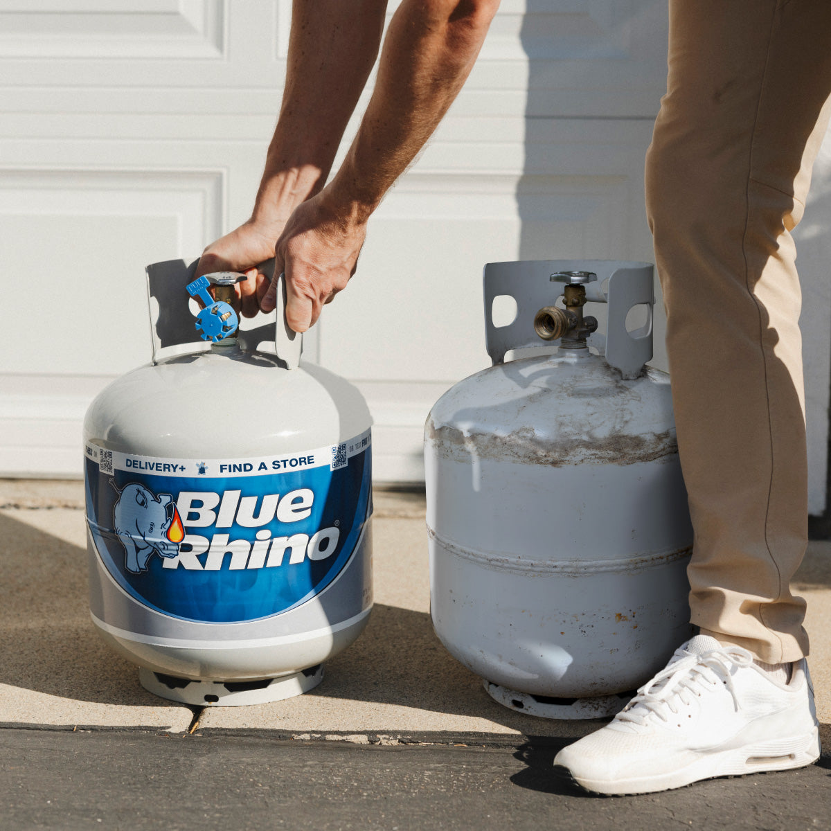 Person holding a Blue Rhino propane tank next to another tank outdoors.