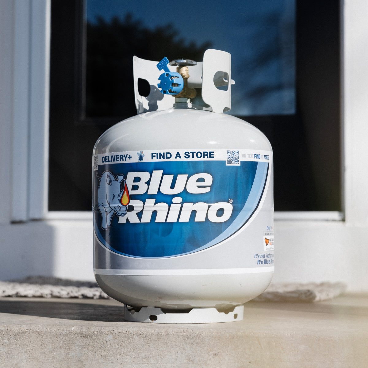 A fresh Blue Rhino propane tank on a concrete surface with a window in the background.