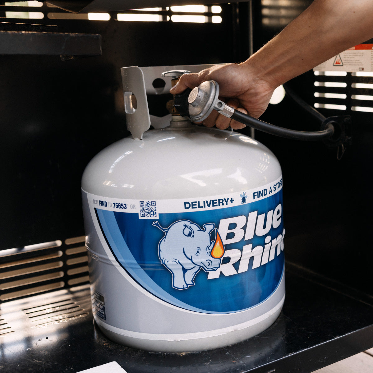 A Person connecting a fresh Blue Rhino propane tank to a standup propane grill in the background.