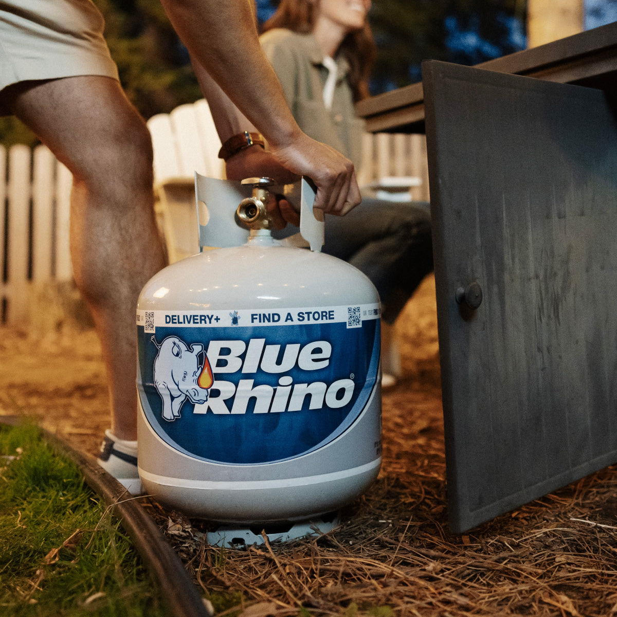 A man connecting a fresh Blue Rhino propane tank to a propane fire pit in the backyard.