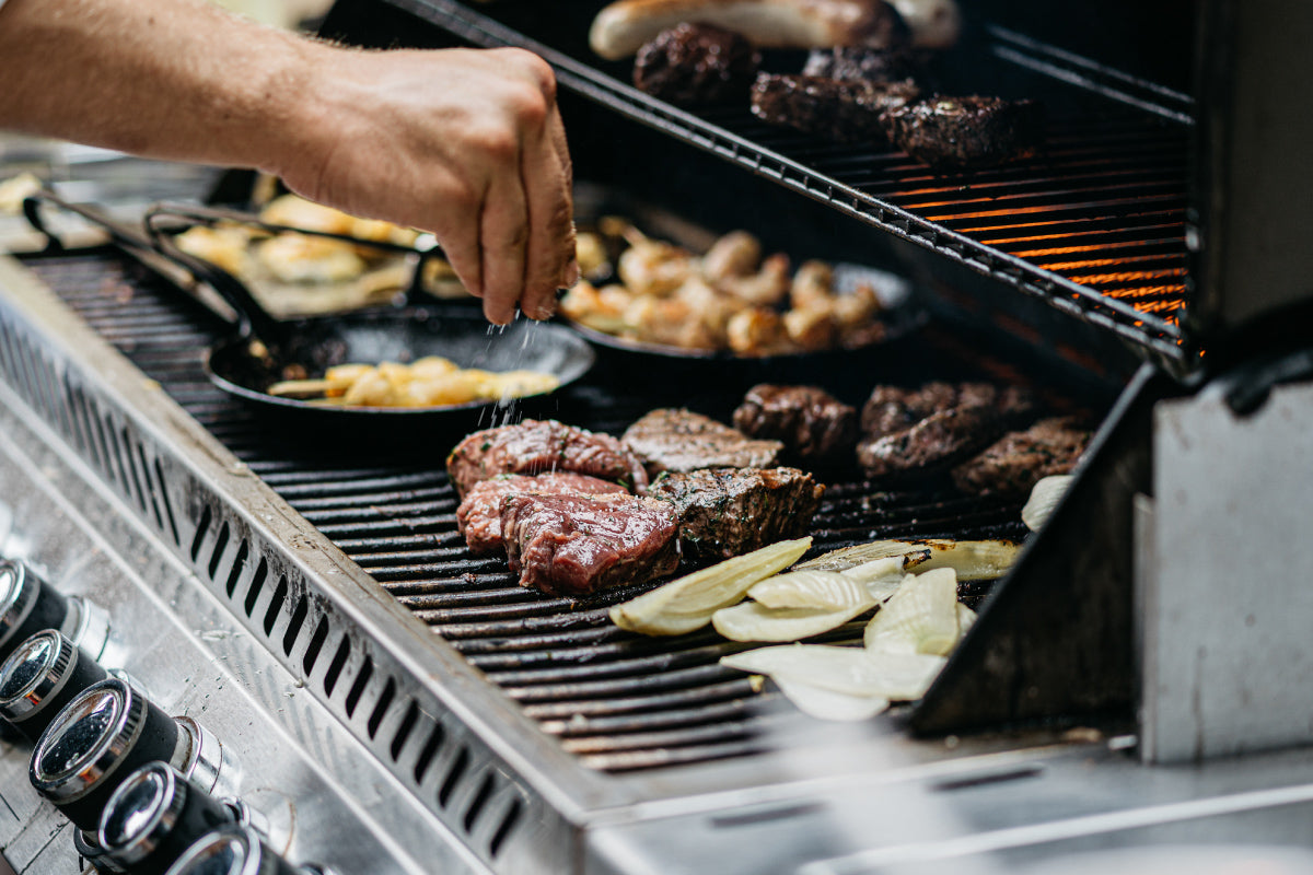A male hand sprinkling seasoning on a steak while grilling other food on a standup propane grill.