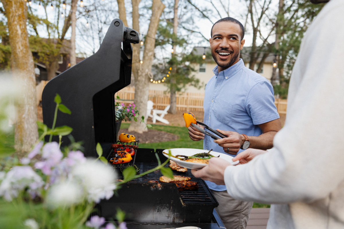 A man using tongs to place a grilled yellow pepper on a plate. Other food on a standup propane grill on the patio in the backyard.